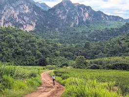 A cyclist on a narrow dirt path winds through vibrant green fields toward dramatic limestone karst mountains.