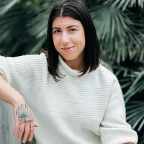 Hannah Deitch in a white sweater leans on a railing, smiling with a tattooed flower on her hand. Green foliage in the background. Serene mood.