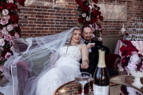 The bride and groom cosy up on a sofa with a drunk in love neon sign behind them