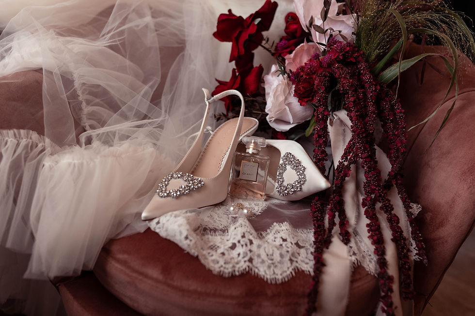Bridal shoes, perfume, veil, and red-white flowers arranged on a blush chair, creating a romantic wedding mood.