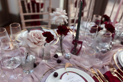 Elegant pink-and-burgundy banquet table with gold-rimmed glassware, candles, roses, and mirrored ornaments in a soft indoor setting.