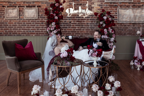 The bride and groom cosy up on a sofa with a drunk in love neon sign behind them and the bride drinks prosecco