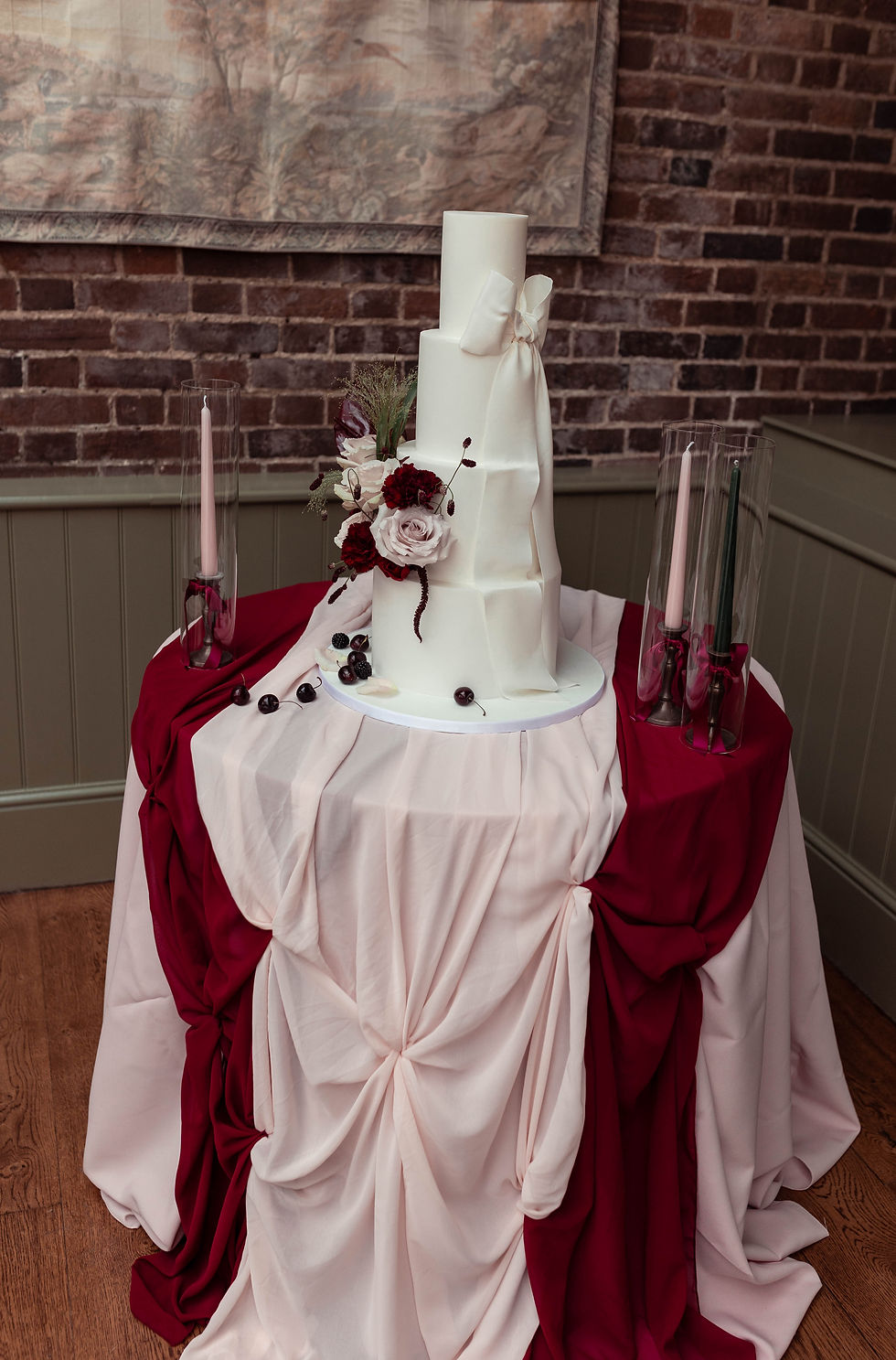Elegant white tiered wedding cake with burgundy and blush draped table, floral accents, and candles against a brick wall.