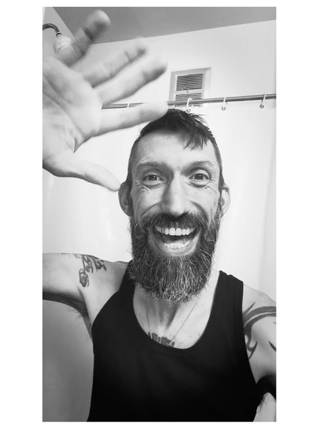 black and white still of jason wyman with their hand up. they are wearing a black tank top. and they are in their bathroom.