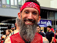 Jason Wyman / Queerly Complex at Folsom Street Fair wearing a red bandanna and a red mesh tank top with a black leather tie. They are smiling with a long graying bear and tattoos. Photo by Mark McBeth