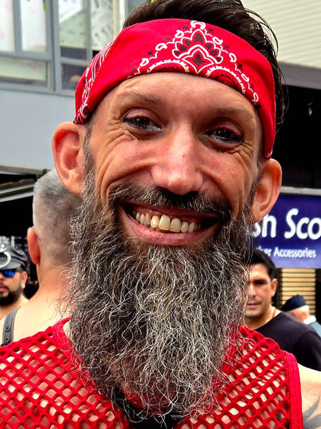 Jason Wyman / Queerly Complex at Folsom Street Fair wearing a red bandanna and a red mesh tank top with a black leather tie. They are smiling with a long graying bear and tattoos. Photo by Mark McBeth