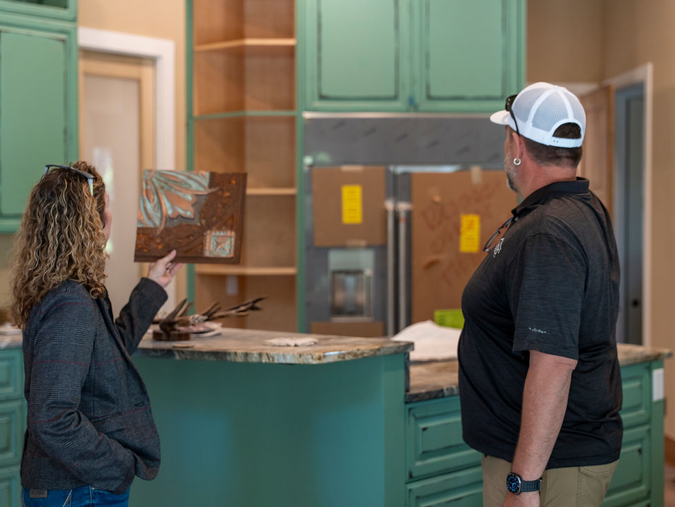Two people in a teal kitchen showroom inspect a decorative wall tile sample, with cabinets and a stainless fridge behind them.