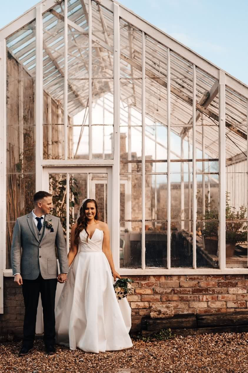 Bride in Suzanne Neville gown and groom in grey suit stand holding hands, smiling in front of a glass greenhouse on a sunny day on grounds of Coombe lodge