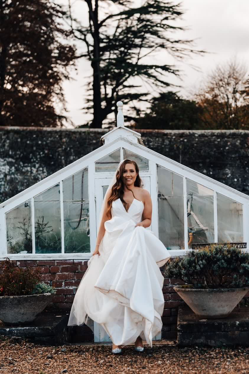 Bride in a Suzanne Neville wedding dress stands smiling in front of a small greenhouse on grounds of Coombe lodge
