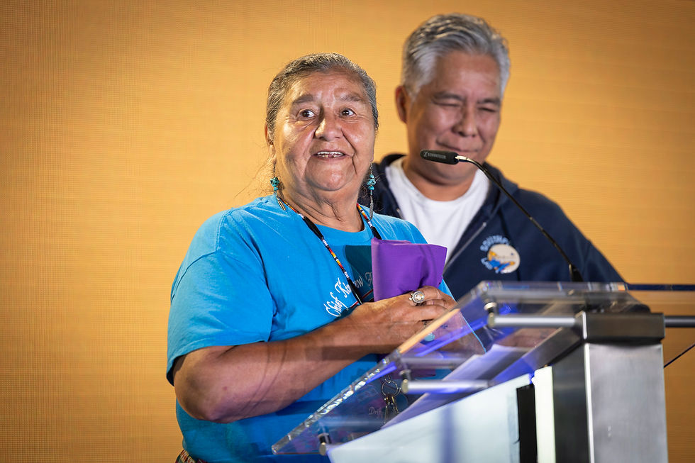 Two people speak at a podium. The woman in a blue shirt holds a purple cloth, smiling. The background is warm yellow.