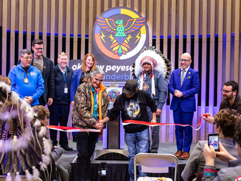 People gathered at a ribbon-cutting ceremony in a modern room, one man in a feathered headdress, screen showing "King Thunderbird Centre".