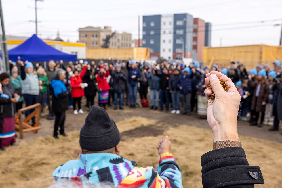 A person in colorful clothing holds tobacco aloft at a ceremony, with a large crowd gathered outdoors under gray skies. Buildings are visible in the background.