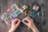 Hands stringing a blue bead with tools and colorful beads scattered on a wooden surface. Emphasizes creativity and crafting.