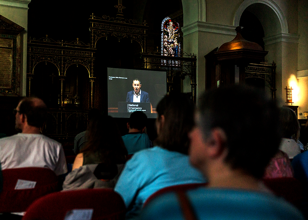 Audience in a dark church watches a projected speaker; screen shows National Emergency and Professor Mike Berners-Lee, with stained glass behind.