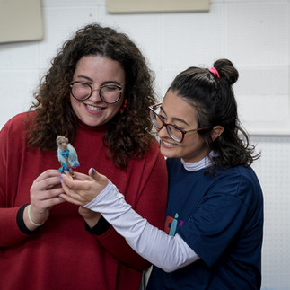 Duas mulheres sorrindo seguram e observam um boneco em miniatura.