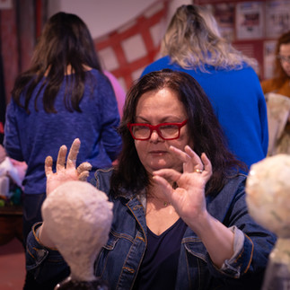 Mulher com óculos fazendo uma escultura de cabeça de papel machê, ao fundo outras pessoas fazem o mesmo.
