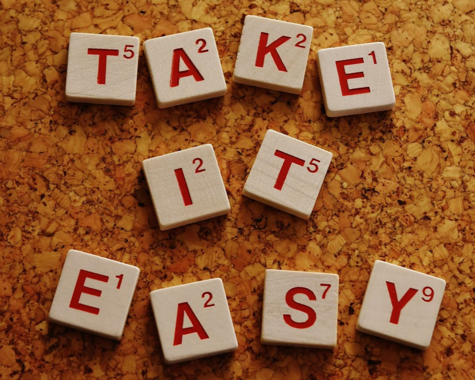 Scrabble tiles spell "TAKE IT EASY" on a cork board. Red letters on white tiles convey a relaxed mood.