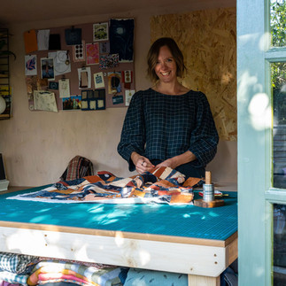 Helen stands behind her cutting table. She smiles at the camera as she pins fabric pieces. On the wall behind her is a mood board displaying fabric swatches, buttons and pictures.