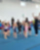 Group photo of 9 young gymnasts and 4 coaches. Everyone is standing on the floor in a group, stretching with their arms up above their heads.