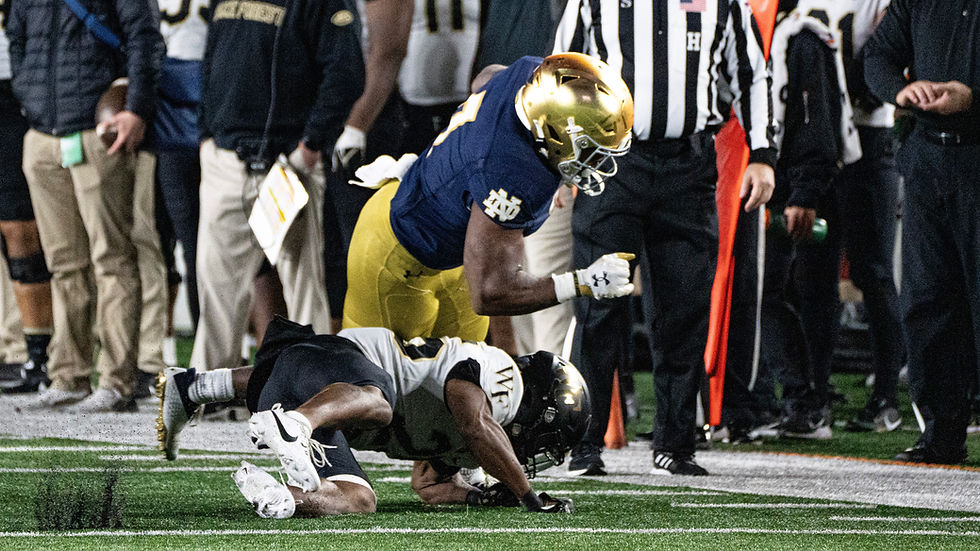 Football player in gold helmet and blue uniform tackles another player in black and white on a football field. Referee in background.