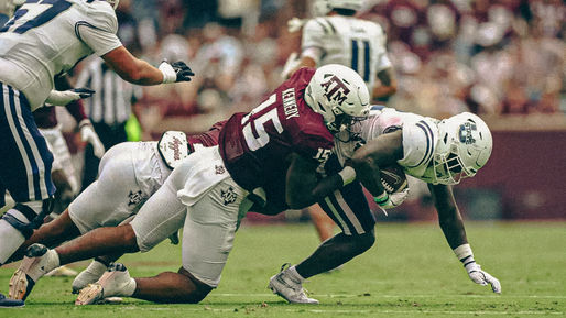 Football player in a maroon jersey tackles an opponent in white, mid-action on the field. Crowd in background. Intense, dynamic scene.