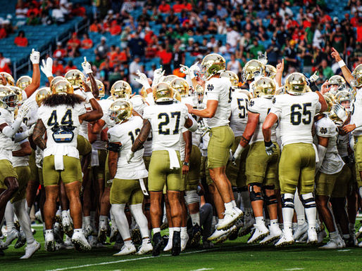 Football team in white and gold gear huddles, raising hands in unity on a green field. Stadium crowd in colorful attire in the background.