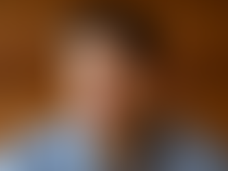 Sam Altman. A man in a blue shirt looks thoughtful against a blurred wooden background. The expression is focused and contemplative. No text present.