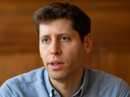 Sam Altman. A man in a blue shirt looks thoughtful against a blurred wooden background. The expression is focused and contemplative. No text present.