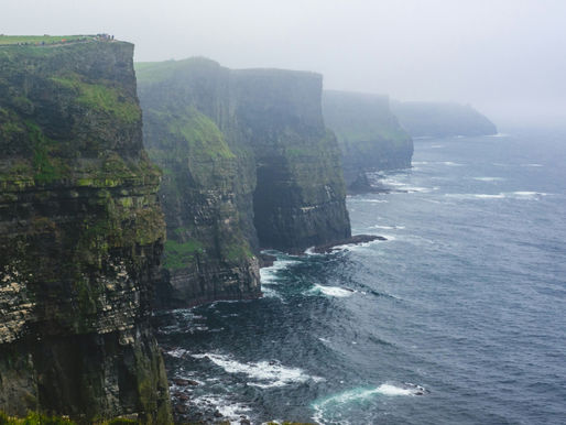 Foggy cliffs meeting the ocean, with lush green tops. Waves crash below. Overcast, moody atmosphere. Small distant figures on cliff edge.