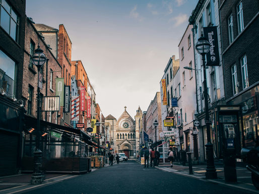 Street view of a busy urban area with historic church in the background. Colorful signs on buildings. People walking. Evening light.