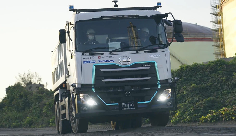 A white UD truck with blue accents drives on a dirt road. Two people in helmets are inside. Factory tanks and greenery in the background.