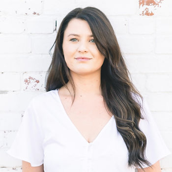 A beautiful woman with long, brown, wavy hair is standing in outside in front of a white painted brick wall.