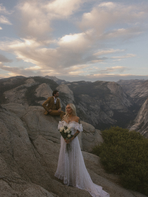 A bride standing off to the side in front of a groom sitting on a rock at their Yosemite elopement