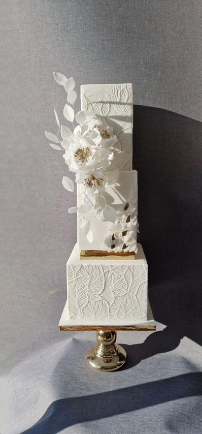 Tall 3-tier square white patterned textured cake with large wafer gold cantered peonies and wafer winding leaves. Gold cake seperators giving extra height.