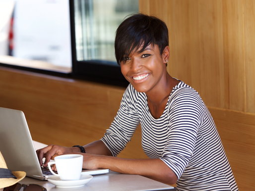 Smiling person in striped shirt using a laptop at a cafe. Wooden walls, a straw hat, and a coffee cup are visible. Relaxed and cheerful mood.