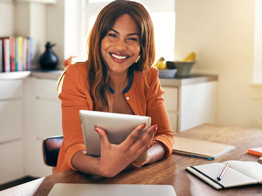 Smiling woman in orange shirt holding a tablet at a wooden desk with notebooks. Bright kitchen setting with books and plants in the background.