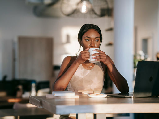 Woman drinking coffee at a wooden table with a laptop, books, and cookies. Sunlit modern room, relaxed mood, wearing a necklace.