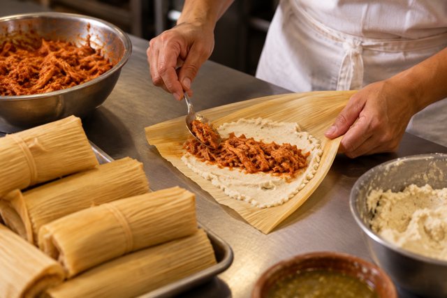 Illustration: Chef assembling corn husk tamales. Fictional scene; not a photograph of this business or its actual menu.