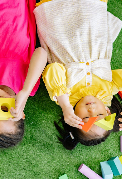Two children lying on green turf and playing with colourful building blocks.