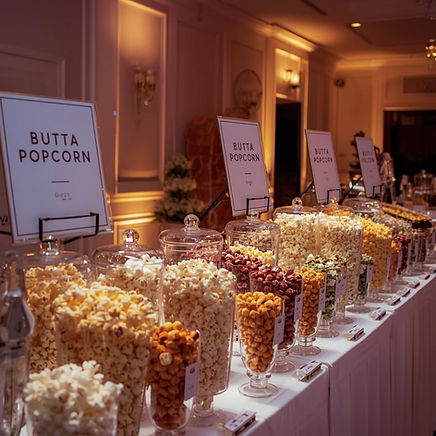 Elegant popcorn bar display featuring multiple glass jars filled with assorted Butta Popcorn flavors, neatly labeled and arranged on a long table at a formal event with warm ambient lighting.