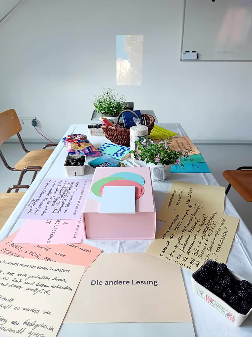 Foto von Tisch, darauf bunte Notizzettel, eine rosa Schachtel, Brombeeren, Snacks, Blümchen, im Hintergrund eine Projektion und eine Tafel, im Vordergrund steht "Die andere Lesung".
