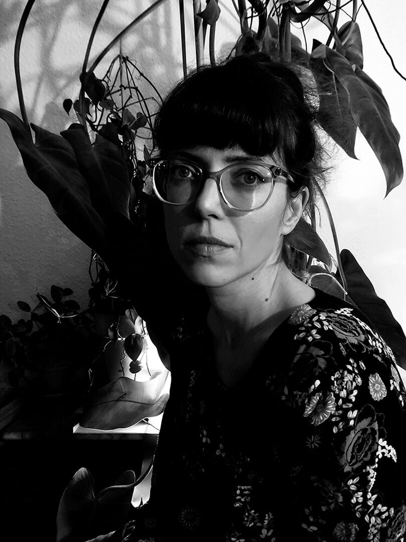 Ayna Steigerwald (Foto privat). Porträt in schwarz-weiß. Ayna sitzt vor einem beleuchteten Hintergrund mit Pflanzen. Sie blickt ernst in die Kamera, trägt die dunklen Haare hochgebunden, eine Brille und ein schwarzes, geblümtes Shirt.