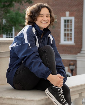 A photograph of the artist, Meo Peltier. She is seated casually on a wall, smiling, with her hands on her knee.