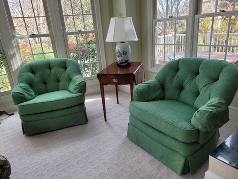 Two matching green upholstered tufted armchairs with skirted bases and matching armcovers, placed on a light patterned carpet in front of large windows, with a side table and lamp between them, showcasing custom upholstered furniture by Mill End Shop.