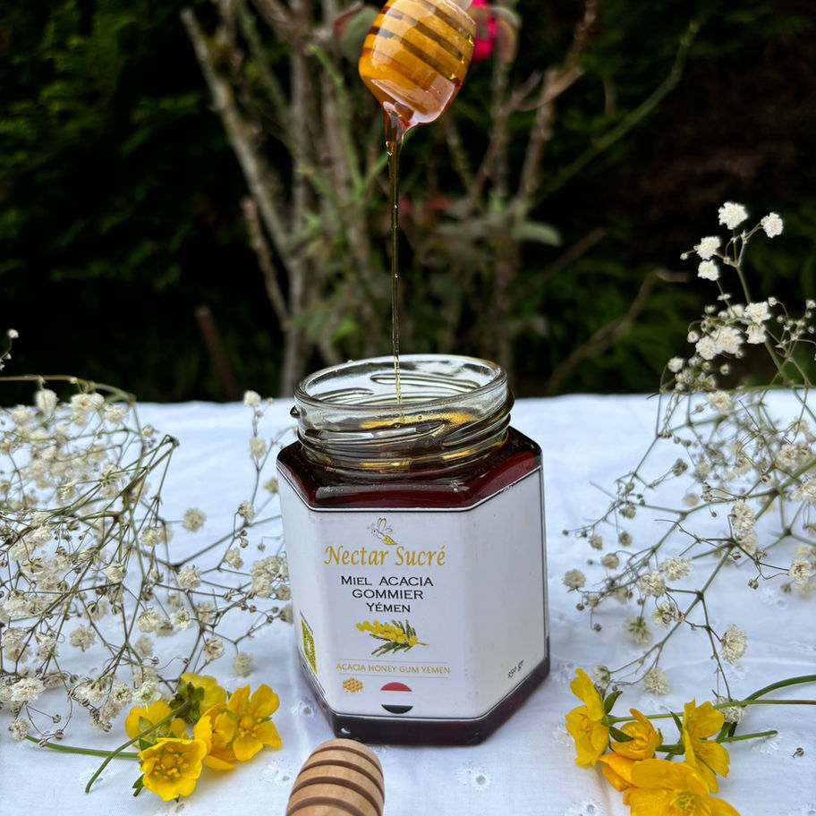 Pot de miel d’acacia gommier du Yémen Nectar Sucré ouvert, miel doré coulant d’une cuillère en bois, décor fleuri naturel.