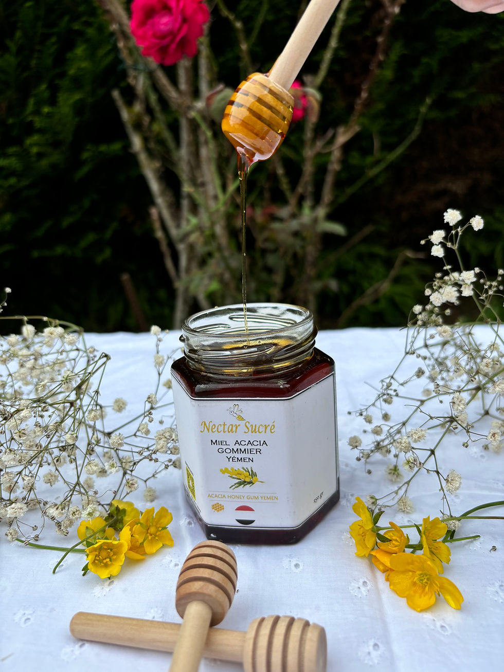 Pot de miel d’acacia gommier du Yémen Nectar Sucré ouvert, miel doré coulant d’une cuillère en bois, décor fleuri naturel.