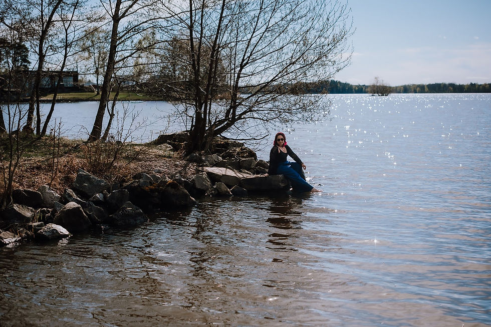 Kvinna sitter på klippor vid en glittrande sjö under en stillsam vårdag med bar natur, mjukt ljus och känsla av närvaro och lugn.