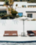 Lounge chairs with white towels beside a swimming pool, shaded by a large umbrella, surrounded by lush greenery and a modern white building.