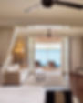 Luxurious hotel room with a beige sofa, wooden table, and open curtains revealing a balcony with ocean views. A serene and inviting atmosphere.