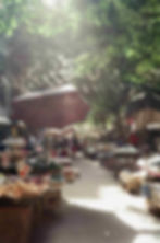 A busy market scene with colourful umbrellas, vendors selling fruits, vegetables, and goods, surrounded by trees.